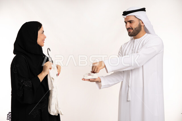 Family atmosphere full of affection, preparing birth supplies, concept of happy and close-knit family, spending enjoyable family time, close-up portrait of an Arab Gulf Emirati family of father and mother carrying clothes for their child, care and attention for children, white background