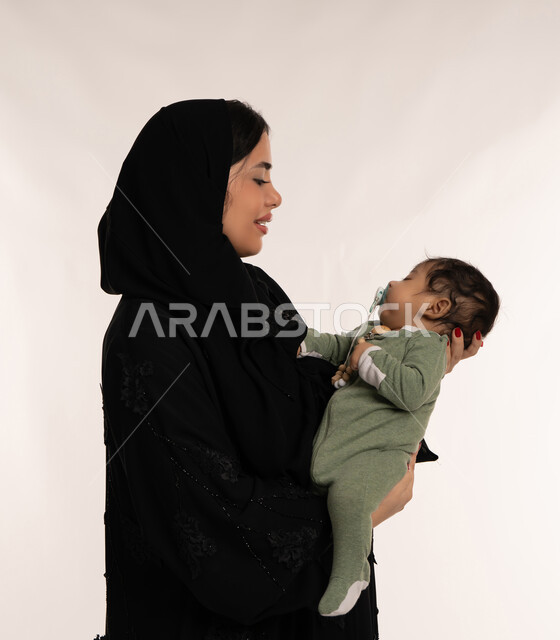 بناء علاقة متينة مع الأبناء، استمداد الرضيع الطمأنينة والاستقرار من الأم، بورتريه مقرب من الجانب لسيدة عربية خليجية إماراتية ترتدي الحجاب والعباءة تحمل طفلها بحنان، تلبية احتياجات الأطفال الجسدية والعاطفية، امرأة سعودية تقوم بقضاء وقت ممتع مع ابنها، خلفية بيضاء