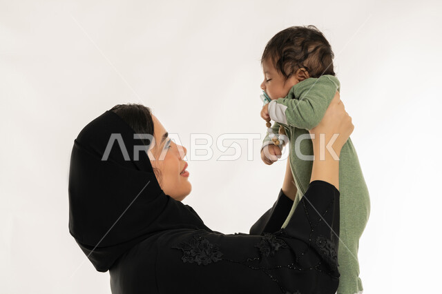 Meeting children's physical and emotional needs, building a strong relationship with children, infant drawing reassurance and stability from mother, close-up side portrait of Emirati Gulf Arab woman wearing hijab and abaya holding her child tenderly, Saudi woman having fun with her son, white background