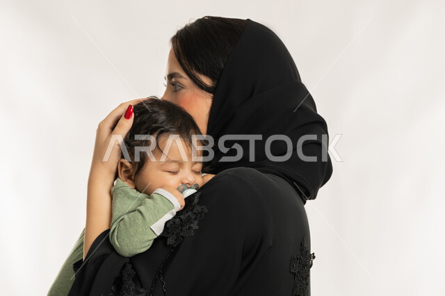 استمداد الرضيع الطمأنينة والاستقرار من الأم، بورتريه مقرب من الجانب لسيدة عربية خليجية إماراتية ترتدي الحجاب والعباءة تحمل طفلها بحنان، تلبية احتياجات الأطفال الجسدية والعاطفية، امرأة سعودية تقوم بقضاء وقت ممتع مع ابنها، بناء علاقة متينة مع الأبناء، خلفية بيضاء