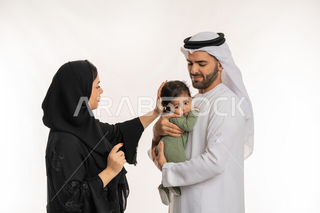 إيماءات الحب والحنان على الصغار, تحمل المسؤولية والعناية بالأطفال، قضاء وقت ممتع مع الطفل, بورتريه مقرب لعائلة عربية خليجية إماراتية مع ابنهم الرضيع, مفهوم الاسرة السعيدة, ملاعبة الأبناء بعطف واهتمام، خلفية بيضاء