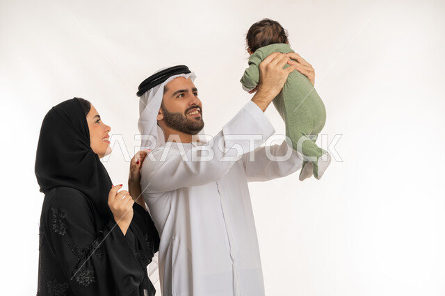ملاعبة الأبناء بعطف واهتمام، قضاء وقت ممتع مع الطفل, بورتريه مقرب لعائلة عربية خليجية إماراتية مع ابنهم الرضيع, إيماءات الحب والحنان على الصغار, تحمل المسؤولية والعناية بالأطفال، مفهوم الاسرة السعيدة, خلفية بيضاء
