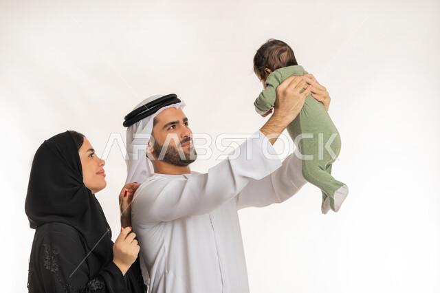 مفهوم الاسرة السعيدة, ملاعبة الأبناء بعطف واهتمام، قضاء وقت ممتع مع الطفل, بورتريه مقرب لعائلة عربية خليجية إماراتية مع ابنهم الرضيع, إيماءات الحب والحنان على الصغار, تحمل المسؤولية والعناية بالأطفال، خلفية بيضاء