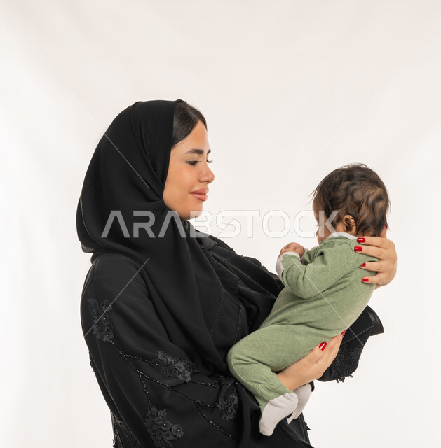 استمداد الرضيع الطمأنينة والاستقرار من الأم، بورتريه مقرب من الجانب لسيدة عربية خليجية إماراتية ترتدي الحجاب والعباءة تحمل طفلها بحنان، تلبية احتياجات الأطفال الجسدية والعاطفية، امرأة سعودية تقوم بقضاء وقت ممتع مع ابنها، بناء علاقة متينة مع الأبناء، خلفية بيضاء