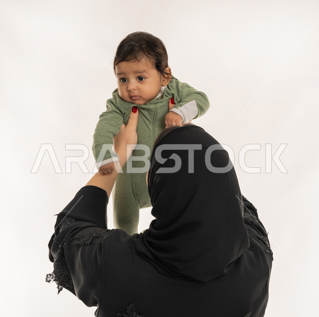 بناء علاقة متينة مع الأبناء، استمداد الرضيع الطمأنينة والاستقرار من الأم، بورتريه مقرب من الخلف لسيدة عربية خليجية إماراتية ترتدي الحجاب والعباءة تحمل طفلها بحنان، تلبية احتياجات الأطفال الجسدية والعاطفية، امرأة سعودية تقوم بقضاء وقت ممتع مع ابنها، خلفية بيضاء