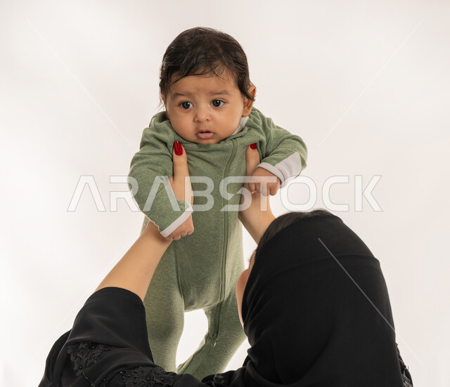 امرأة سعودية تقوم بقضاء وقت ممتع مع ابنها، بناء علاقة متينة مع الأبناء، استمداد الرضيع الطمأنينة والاستقرار من الأم، بورتريه مقرب من الخلف لسيدة عربية خليجية إماراتية ترتدي الحجاب والعباءة تحمل طفلها بحنان، تلبية احتياجات الأطفال الجسدية والعاطفية، خلفية بيضاء