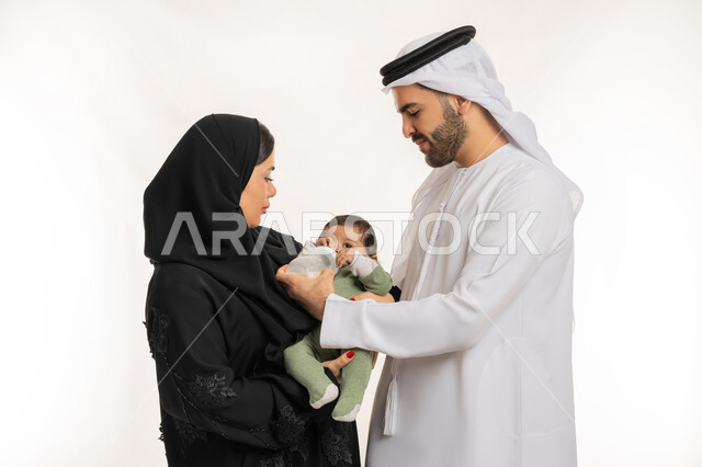 أجواء أسرية مليئة بالمودة, مفهوم الأسرة السعيدة المترابطة, قضاء وقت عائلي ممتع، بورتريه مقرب لعائلة عربية خليجية إماراتية يقومون بإعطاء طفلهما الرضيع الحليب الصناعي من عبوة الرضاعة, الرعاية والاهتمام بالأطفال، الأمان و العطف على الأبناء، خلفية بيضاء