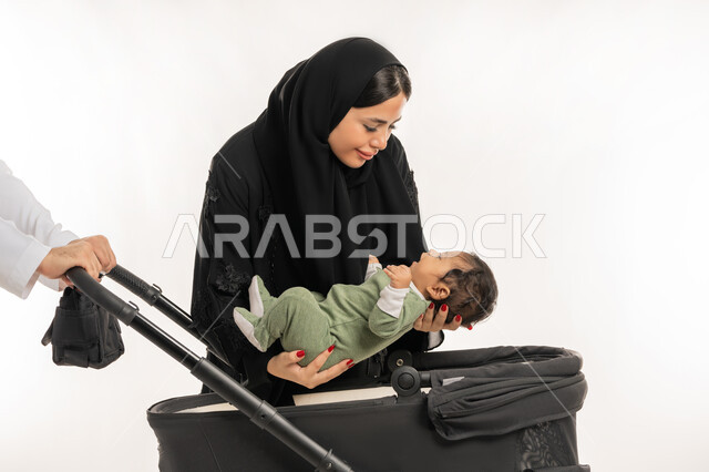 أجواء أسرية مليئة بالمودة، مفهوم الأسرة السعيدة المترابطة, بورتريه مقرب لعائلة عربية خليجية إماراتية من أب وأم يضعان ابنهما الرضيع في عربة الطفل, قضاء وقت ممتع مع الأطفال في الخارج، رحلة وزيارة عائلية ممتعة، الرعاية والاهتمام بالأبناء، خلفية بيضاء