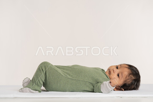 رضيع سعودي يقوم بالنظر إلى شيء ما بإيماءات الدفء والهناء, الملامح الناعمة والبريئة، بورتريه لطفل عربي خليجي إماراتي لطيف يرتدي لباس قطني مستلقي على ظهره فوق بطانية, مفهوم البراءة والطفولة, خلفية بيضاء
