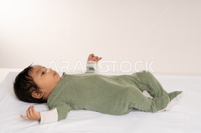 رضيع سعودي يقوم بالنظر إلى شيء ما بإيماءات الدفء والهناء, الملامح الناعمة والبريئة، بورتريه لطفل عربي خليجي إماراتي لطيف يرتدي لباس قطني مستلقي على ظهره فوق بطانية, مفهوم البراءة والطفولة, خلفية بيضاء