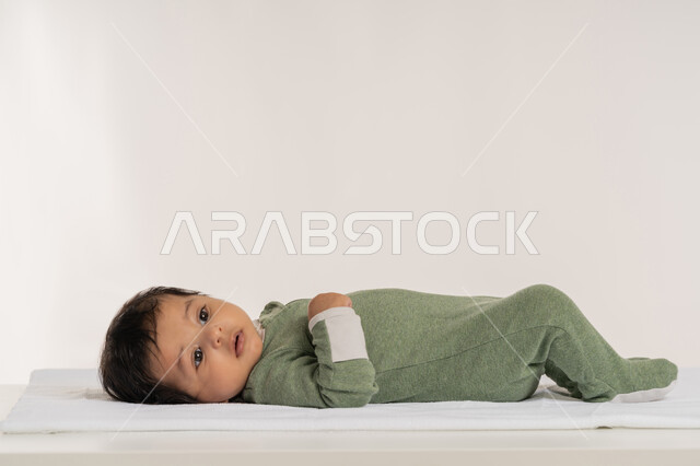 الملامح الناعمة والبريئة، بورتريه لطفل عربي خليجي إماراتي لطيف يرتدي لباس قطني مستلقي على ظهره فوق بطانية, رضيع سعودي يقوم بالنظر إلى شيء ما بإيماءات الدفء والهناء, مفهوم البراءة والطفولة, خلفية بيضاء