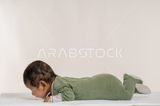 رضيع سعودي يقوم بالنظر إلى شيء ما بإيماءات الدفء والهناء, الملامح الناعمة والبريئة، بورتريه لطفل عربي خليجي إماراتي لطيف يرتدي لباس قطني مستلقي على بطنه فوق بطانية, مفهوم البراءة والطفولة, خلفية بيضاء