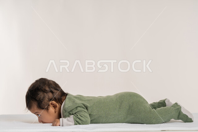 الملامح الناعمة والبريئة، بورتريه لطفل عربي خليجي إماراتي لطيف يرتدي لباس قطني مستلقي على بطنه فوق بطانية, مفهوم البراءة والطفولة, رضيع سعودي يقوم بالنظر إلى شيء ما بإيماءات الدفء والهناء, خلفية بيضاء