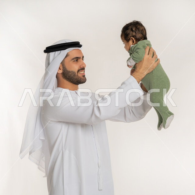 تحمل المسؤولية والعناية بالأطفال، بورتريه مقرب من الجانب لأب عربي خليجي إماراتي يرتدي الكندورة والغترة يحمل ابنه الرضيع بيديه, قضاء وقت ممتع مع الطفل, إيماءات الحب والحنان على الصغار, ملاعبة الأبناء بعطف واهتمام، خلفية بيضاء