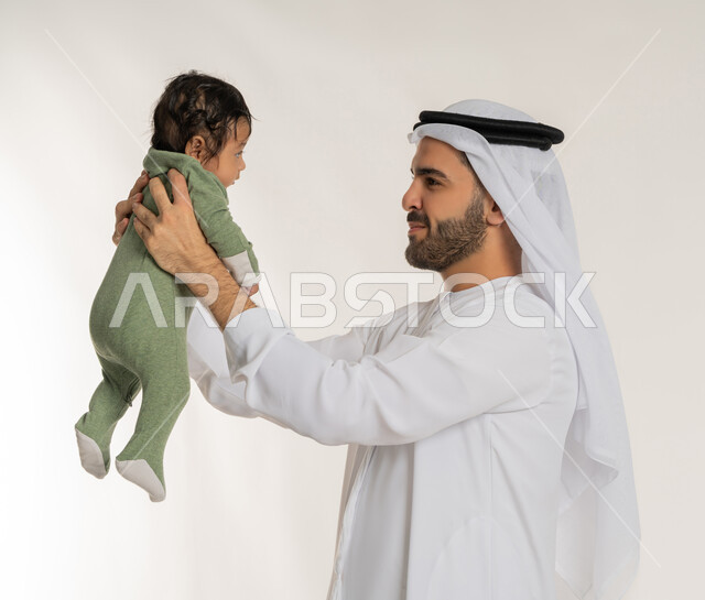 إيماءات الحب والحنان على الصغار, تحمل المسؤولية والعناية بالأطفال، بورتريه مقرب من الجانب لأب عربي خليجي إماراتي يرتدي الكندورة والغترة يحمل ابنه الرضيع بيديه, قضاء وقت ممتع مع الطفل, ملاعبة الأبناء بعطف واهتمام، خلفية بيضاء