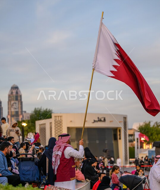 الاحتفال بالأعياد والمناسبات الوطنية، الفخر والاعتزاز بالهوية القطرية, اليوم الوطني لدولة قطر 18 ديسمبر, مفهوم الحب والانتماء للبلاد, صورة من الخلف لرجل عربي خليجي قطري يرتدي الثوب التقليدي والشماغ يحمل العلم القطري إحدى فعاليات الدوحة