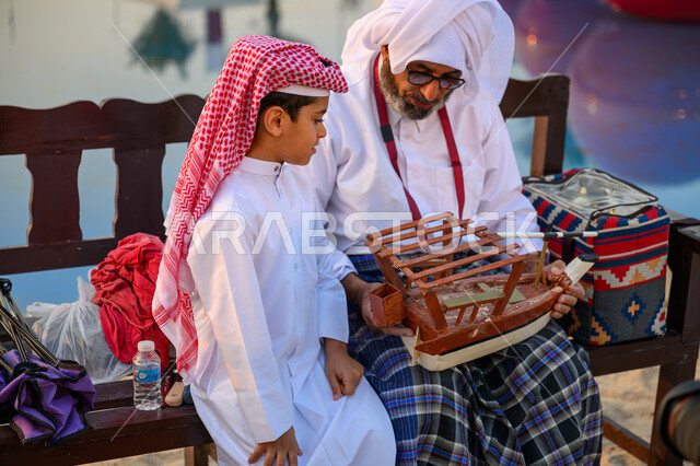 الاحتفال بالأعياد والمناسبات الوطنية, ذكرى باليوم الوطني لدولة قطر 18 ديسمبر, فعالية ونشاط ترفيهي احتفالي, صياد عربي خليجي قطري يرتدي الثوب التقليدي يجلس مع ابنه ويمسك بيده مجسم قارب صيد خشبي صغير, مهرجان كتارا للقوارب الشراعية التقليدية, تراث الأجداد القطري