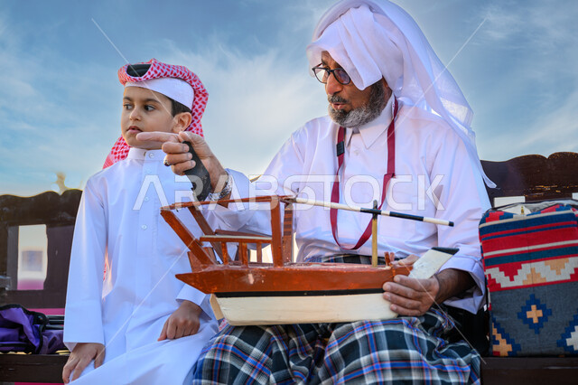 الاحتفال بالأعياد والمناسبات الوطنية, ذكرى اليوم الوطني لدولة قطر 18 ديسمبر, صياد عربي خليجي قطري يرتدي الثوب التقليدي يجلس مع ابنه ويمسك بيده مجسم قارب صيد خشبي صغير ويشير بإصبع السبابة لشيء ما, مهرجان كتارا للقوارب الشراعية التقليدية, تراث الأجداد القطري