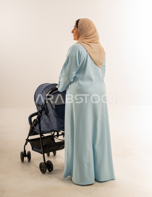 Meeting children's physical and emotional needs, taking a walk and tour with mother, portrait from behind of an Arab Gulf Omani woman wearing hijab and abaya pulling a baby stroller, building a strong relationship with children, Saudi woman having fun with her son, full body, white background