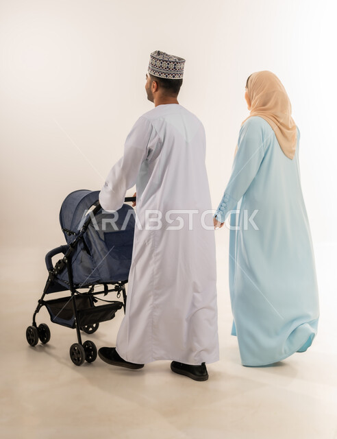 أهمية الأهل ودورهم الأساسي في تربية ونشأة الأبناء, بورتريه من الخلف لرجل عربي خليجي عماني يرتدي الدشداشة والكمة يقف مع زوجته يقوم بجر عربة الطفل، القيام بنزهة وجولة عائلية، الرعاية والاهتمام بالأولاد, عائلة عمانية تقضي وقت ممتع، الجسم بالطول الكامل، خلفية بيضاء