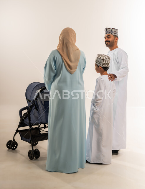 الرعاية والاهتمام بالأولاد, عائلة عمانية تقضي وقت ممتع، أهمية الأهل ودورهم الأساسي في تربية الأبناء, بورتريه من الخلف لسيدة عربية خليجية عمانية ترتدي الحجاب والعباءة تقف مع زوجها وابنها تقوم بجر عربة الطفل، القيام بجولة عائلية، الجسم بالطول الكامل، خلفية بيضاء