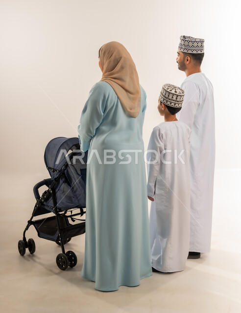 عائلة عمانية تقضي وقت ممتع، أهمية الأهل في تربية الأبناء, بورتريه من الخلف لسيدة عربية خليجية عمانية ترتدي الحجاب والعباءة تقف مع زوجها وابنها تقوم بجر عربة الطفل، القيام بنزهة وجولة عائلية، الرعاية والاهتمام بالأولاد, الجسم بالطول الكامل، خلفية بيضاء