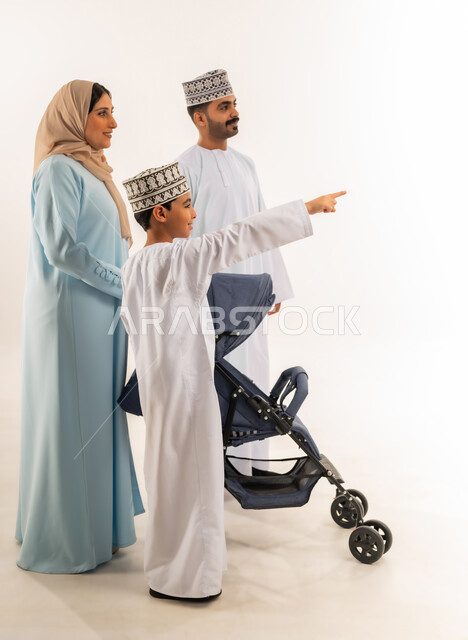 ولد عماني يشير بإصبع السبابة إلى شيء ما, بورتريه من الجانب لسيدة عربية خليجية عمانية ترتدي الحجاب والعباءة تقف مع زوجها وابنها تجر عربة الطفل، القيام بنزهة وجولة عائلية، الرعاية والاهتمام بالأولاد, عائلة عمانية تقضي وقت ممتع، الجسم بالطول الكامل، خلفية بيضاء