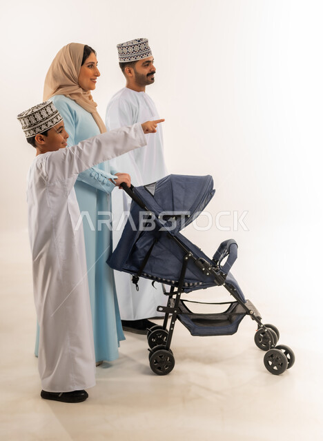 Omani family having fun, Omani boy pointing index finger at something, side portrait of Omani Gulf Arab woman wearing hijab and abaya standing with her husband and son pulling a stroller, family outing, care and attention for children, full body, white background