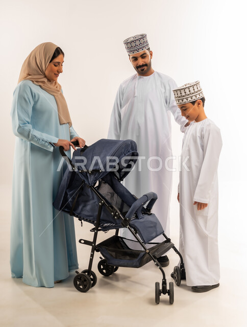 أهمية الأهل ودورهم الأساسي في تربية ونشأة الأبناء, بورتريه لسيدة عربية خليجية عمانية ترتدي الحجاب والعباءة تقف مع زوجها وابنها تقوم بجر عربة الطفل، القيام بجولة عائلية، الرعاية والاهتمام بالأولاد, عائلة عمانية تقضي وقت ممتع، الجسم بالطول الكامل، خلفية بيضاء