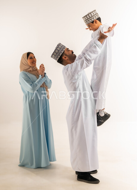 ملاعبة الأبناء وملاطفتهم, بناء علاقة متينة مع الأولاد، مفهوم الأسرة السعيدة المترابطة، بورتريه لرجل عربي خليجي عماني يرتدي الدشداشة والكمة يقف مع زوجته يقوم بحمل ابنه، عائلة عمانية تقضي وقت ممتع، أجواء أسرية مليئة بالمودة، الجسم بالطول الكامل، خلفية بيضاء