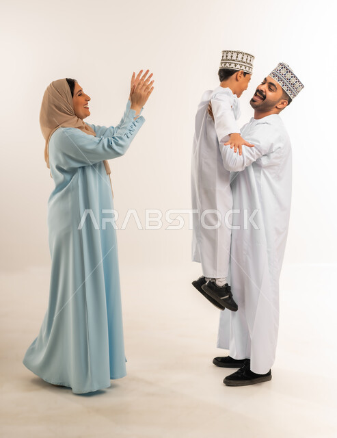 أجواء أسرية مليئة بالمودة، مفهوم الأسرة السعيدة المترابطة، بورتريه لرجل عربي خليجي عماني يرتدي الدشداشة والكمة يقف مع زوجته يقوم بحمل ابنه، عائلة عمانية تقضي وقت ممتع، ملاعبة الأبناء وملاطفتهم, بناء علاقة متينة مع الأولاد، الجسم بالطول الكامل، خلفية بيضاء