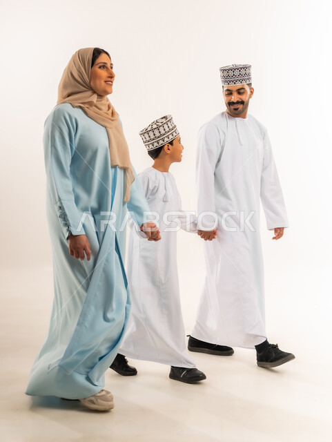 بناء علاقة متينة مع الأولاد، مفهوم الأسرة السعيدة المترابطة، بورتريه لرجل عربي خليجي عماني يرتدي الدشداشة والكمة يمشي مع زوجته وابنه إلى مكان ما، أجواء أسرية مليئة بالمودة، عائلة عمانية تقضي وقت ممتع، الرعاية والاهتمام بالأبناء, الجسم بالطول الكامل، خلفية بيضاء
