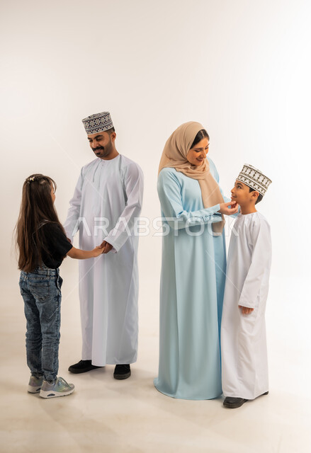 تبادل الحوار والنقاش المسلي، أجواء أسرية مليئة بالمودة، عائلة عمانية تقضي وقت ممتع، مفهوم الأسرة السعيدة المترابطة، بورتريه لرجل عربي خليجي عماني يرتدي الدشداشة والكمة يقف مع زوجته وأبناءه، الرعاية والاهتمام بالأبناء, الجسم بالطول الكامل، خلفية بيضاء