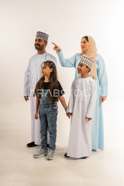 Family atmosphere full of affection, Omani woman pointing index finger at something above, Omani family having fun, concept of happy and close-knit family, portrait of an Arab Gulf Omani man wearing a dishdasha and a kummah standing with his wife and children, full body, white background