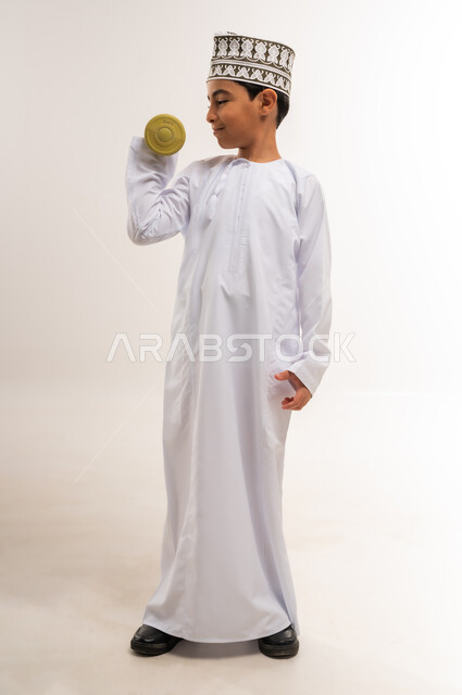 التدرب على رفع الأثقال والقيام بالتمارين الرياضية، بورتريه لفتى عربي خليجي عماني يرتدي الدشداشة والكمة يقوم برفع الدامبلز، اتباع أسلوب حياة صحي، ممارسة الرياضة وبناء الكتلة العضلية، الجسم بالطول الكامل، خلفية بيضاء