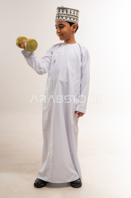 التدرب على رفع الأثقال والقيام بالتمارين الرياضية، بورتريه لفتى عربي خليجي عماني يرتدي الدشداشة والكمة يقوم برفع الدامبلز، اتباع أسلوب حياة صحي، ممارسة الرياضة وبناء الكتلة العضلية، الجسم بالطول الكامل، خلفية بيضاء