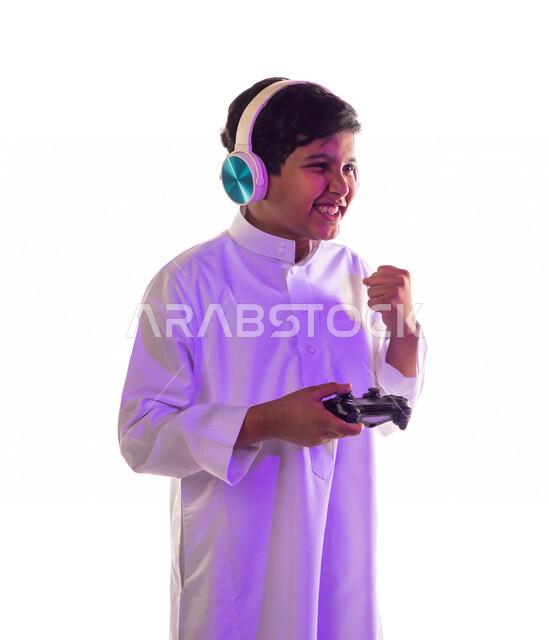 بورتريه لطفل عربي خليجي سعودي مبتسم ، يرتدي سماعة رأس و يمسك بيده عصا التحكم و يلعب بألعاب الفيديو ، بإيماءات و تعابير تدل على الفوز ، المتعة و التسلية بألعاب الفيديو ، ألعاب البلايستيشن الترفيهية ، تقنيات و ألعاب
