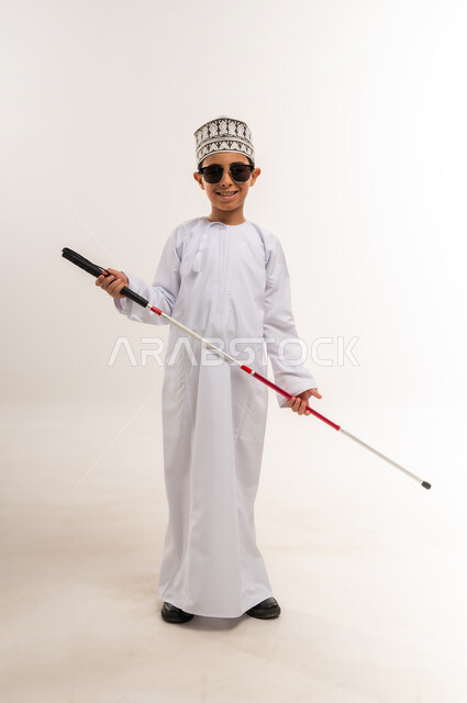 اعتماد الشخص الضرير على العصا البيضاء بشكل كامل في جميع تحركاته, مفهوم الصحة والمرض, بورتريه لفتى عربي خليجي عماني كفيف يرتدي الدشداشة والكمة يضع النظارة الشمسية يحمل بيده العكاز، فقدان الرؤية والبصر, الجسم بالطول الكامل، خلفية بيضاء