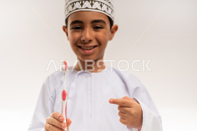المحافظة على ابتسامة ناصعة البياض، بورتريه مقرب لفتى عربي خليجي عماني يرتدي الدشداشة والكمة يمسك بيده الفرشاة يشير إليها, مفهوم العادات الصحية والعناية بصحة الفم، حماية السن من التسوس، التأكد من نظافة الأسنان بعد تناول الطعام، خلفية بيضاء