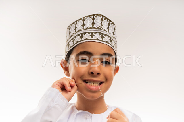 الاهتمام بالأناقة والمظهر الخارجي، مفهوم العناية بصحة الفم، التأكد من نظافة الأسنان بعد تناول الطعام، المحافظة على ابتسامة ناصعة البياض، بورتريه مقرب لفتى عربي خليجي عماني يرتدي الدشداشة والكمة يقوم بتنظيف أسنانه باستخدام الخيط السني, خلفية بيضاء