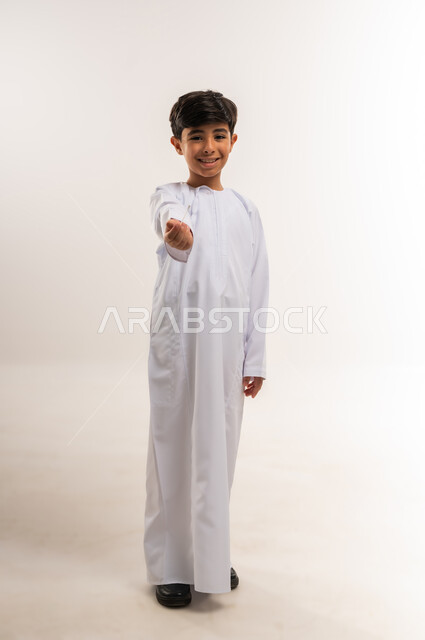 حمل وإمساك شيء ما، مفهوم الاناقة والاهتمام بالمظهر الخارجي, بورتريه لولد عربي خليجي عماني يرتدي الدشداشة و الكمة يرفع يده ينظر إلى الكاميرا بإيماءات السرور والسعادة، صورة الجسم بالطول الكامل, خلفية بيضاء