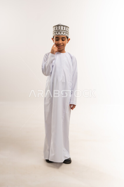 الاهتمام بالمظهر الخارجي، دهن وترطيب الوجه، بورتريه لفتى عربي خليجي عماني يرتدي الدشداشة والكمة يرفع إصبع السبابة يقوم بوضع شيء ما على خده، الجسم بالطول الكامل، خلفية بيضاء