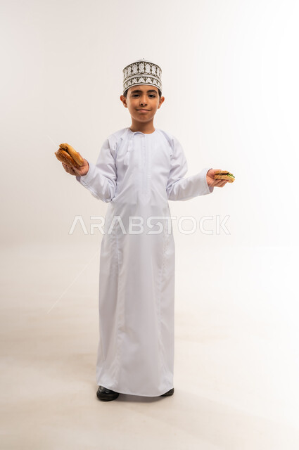 تناول المخبوزات الطازجة الشهية، الحيرة بين الطعام الصحي والضار، الشعور بالجوع والاستعداد لتناول الغداء، بورتريه لولد عربي خليجي عماني يرتدي الدشداشة والكمة يمسك بيديه شطيرة وساندوتش من البرجر ينظر إلى الكاميرا بإيماءات التردد، الجسم بالطول الكامل، خلفية بيضاء