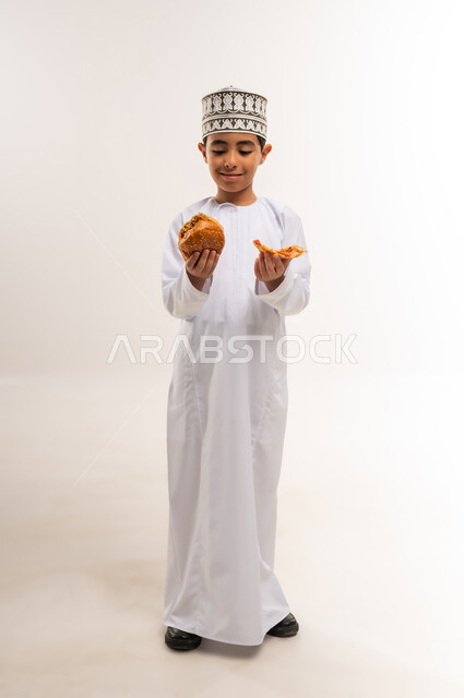 الحيرة بين الطعام الصحي والضار، الشعور بالجوع والاستعداد لتناول الغداء، بورتريه لولد عربي خليجي عماني يرتدي الدشداشة والكمة يمسك بيديه شطيرة وساندوتش من البرجر ينظر إليهما بإيماءات التردد، تناول المخبوزات الطازجة الشهية، الجسم بالطول الكامل، خلفية بيضاء