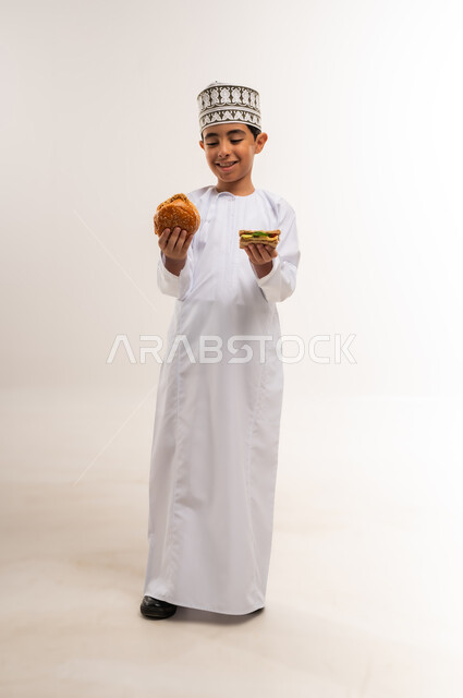 الشعور بالجوع والاستعداد لتناول الغداء، بورتريه لولد عربي خليجي عماني يرتدي الدشداشة والكمة يمسك بيديه شطيرة وساندوتش من البرجر ينظر إليهما بإيماءات التردد، الحيرة بين الطعام الصحي والضار، تناول المخبوزات الطازجة الشهية، الجسم بالطول الكامل، خلفية بيضاء
