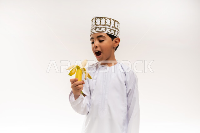 أهمية العناصر الغذائية الموجودة في الموز, الطعام الصحي والمتوازن, بورتريه مقرب لولد عربي خليجي عماني يرتدي الدشداشة والكمة يحمل بيده موزة يقربها إلى فمه، فاكهة طبيعية وطازجة, وجبة مدرسية خفيفة, اتباع نظام غذائي، خلفية بيضاء
