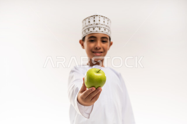 أهمية العناصر الغذائية الموجودة في التفاح, وجبة مدرسية خفيفة, اتباع نظام غذائي، الطعام الصحي والمتوازن, بورتريه مقرب لولد عربي خليجي عماني يرتدي الدشداشة والكمة يحمل بيده تفاحة ينظر إلى الكاميرا بإيماءات السرور، فاكهة طبيعية وطازجة, خلفية بيضاء
