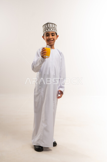 المحافظة على سلامة الجهاز المناعي، فاكهة منعشة ولذيذة، اتباع نظام غذائي صحي، بورتريه لولد عربي خليجي عماني يرتدي الدشداشة والكمة يحمل بيده كوب من عصير البرتقال الطازج، مفهوم العناية والاهتمام بالصحة, الجسم بالطول الكامل، خلفية بيضاء