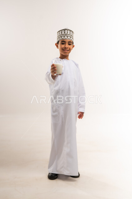 The multiple benefits of dairy products for the body and bones, portrait of an Arab Gulf Omani boy wearing a dishdasha and a kummah holding a cup of milk in his hand, maintaining a healthy immune system, following a healthy diet, the concept of care and attention to health, full-length body, white background