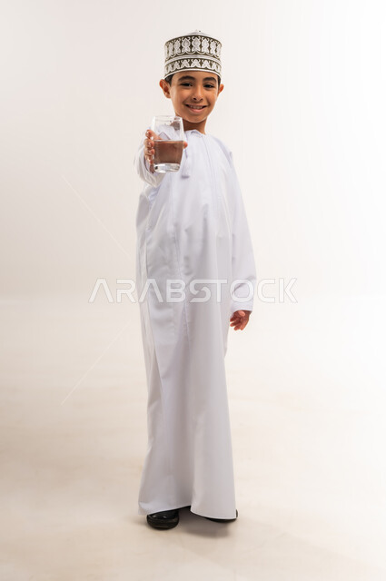 المحافظة على سلامة الجهاز المناعي، التخلص من سموم الجسم، الحفاظ على صحة الكلى ومعالجتها، بورتريه لولد عربي خليجي عماني يرتدي الدشداشة والكمة يحمل بيده كأس من المياه، أهمية شرب الماء النقي للحصول على بشرة نضرة، الجسم بالطول الكامل، خلفية بيضاء